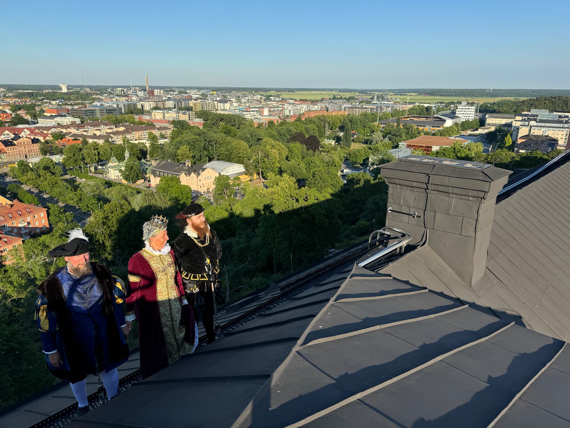 Tre historiska personer i 1500-talsdräkter och takselar står vid en skorsten på Uppsala slotts tak med utsikt över staden
